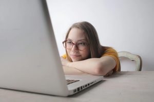 A young woman wearing eyeglasses relaxes at home, leaning on a table while using a laptop.