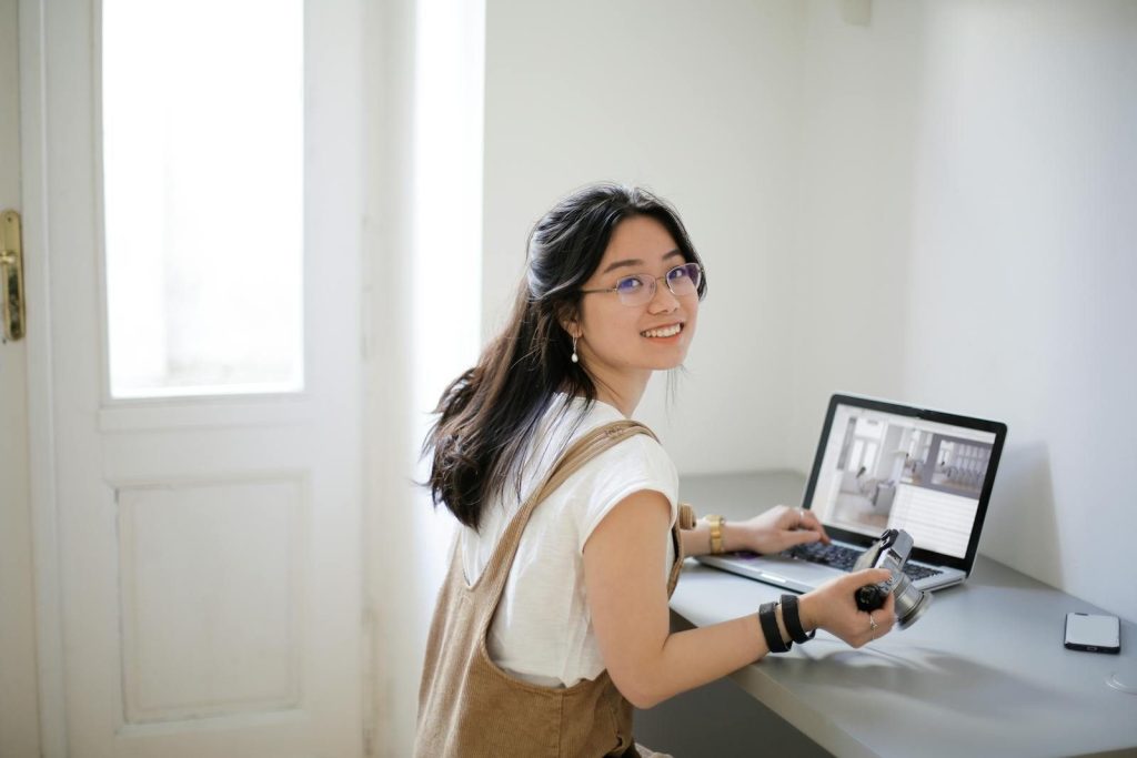 Young woman photographer smiling at desk with laptop indoors, embracing creative work.