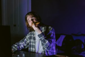 A young man enjoys gaming with headphones while eating pizza in a dimly lit room.