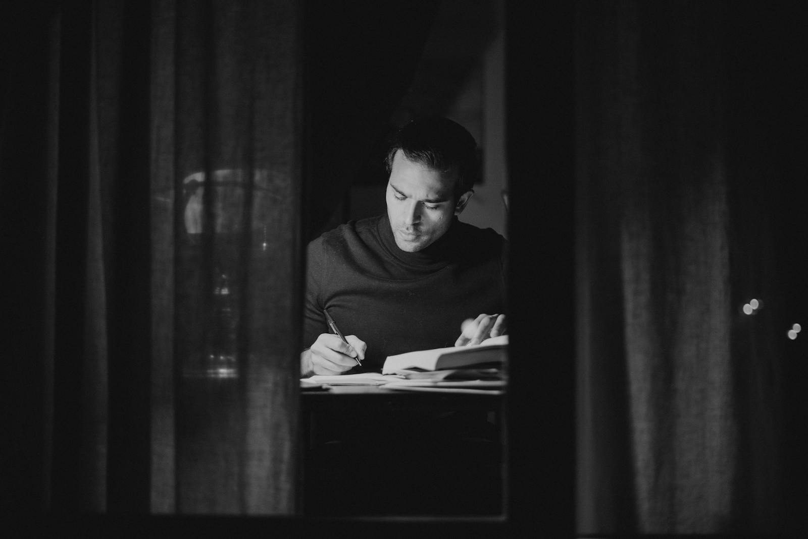 A man deeply focused on studying at night, captured in black and white from an indoor perspective.