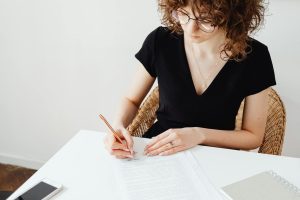 A woman in glasses and black top writing on documents at a white table, focusing intently.
