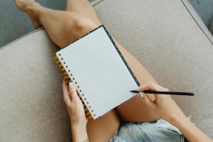 Top view of a woman in denim shorts writing in a notebook with a pen, focusing on relaxation and creativity.