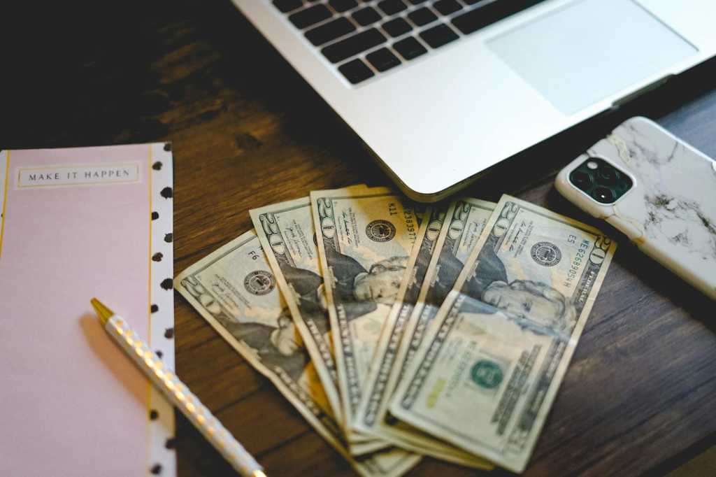 A workspace setup with dollar bills, notepad, and laptop on a wooden desk.