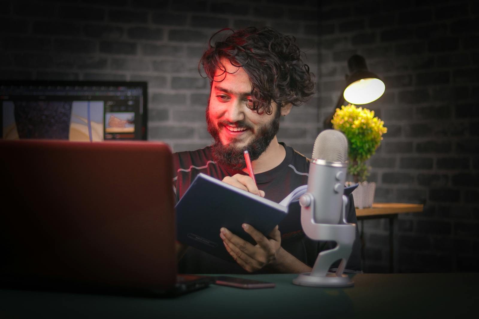 A bearded man writes notes in a cozy home office with a laptop and microphone.