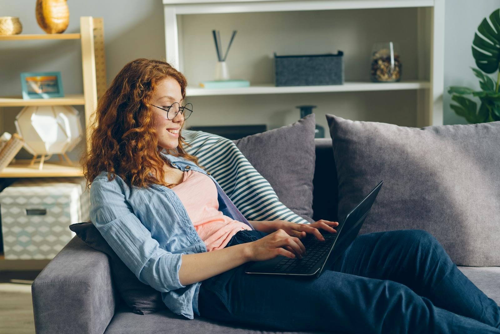 Red-haired woman smiling while working on a laptop comfortably at home.