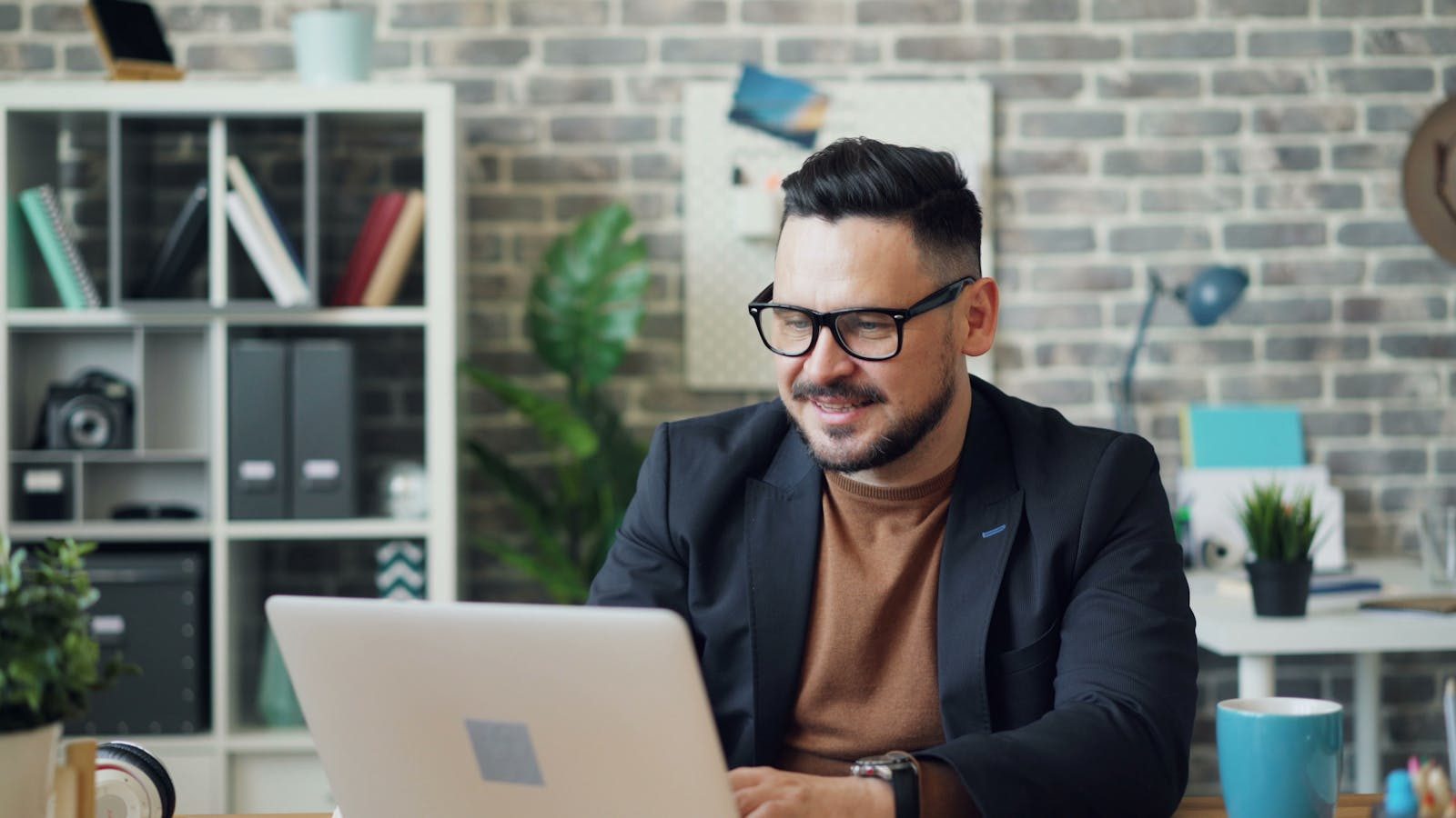 A cheerful man in a casual office setting working on a laptop, wearing eyeglasses and a jacket.