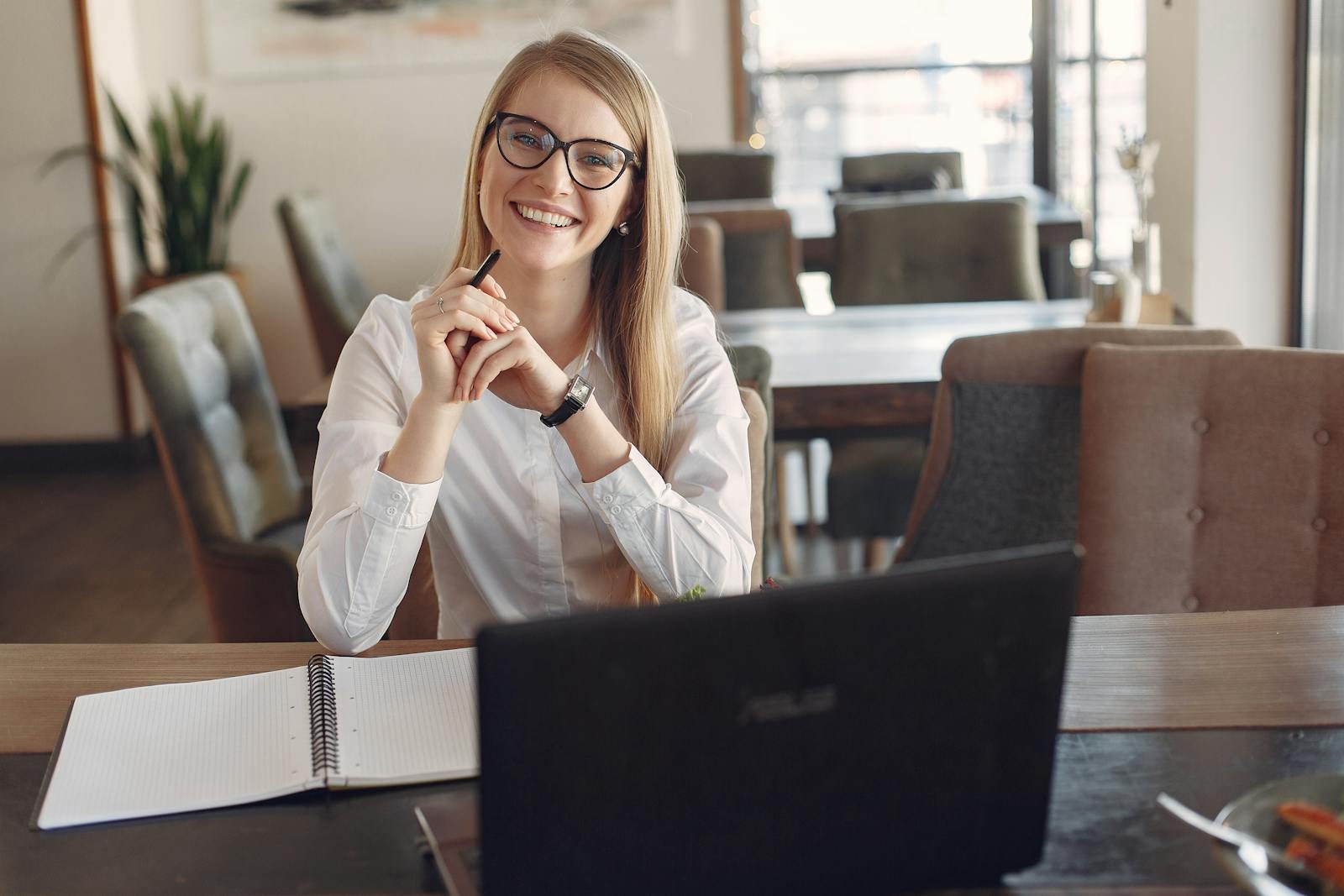 Happy female freelancer wearing white formal shirt sitting at table with laptop and copybook working on business project in cafe and smiling looking at camera