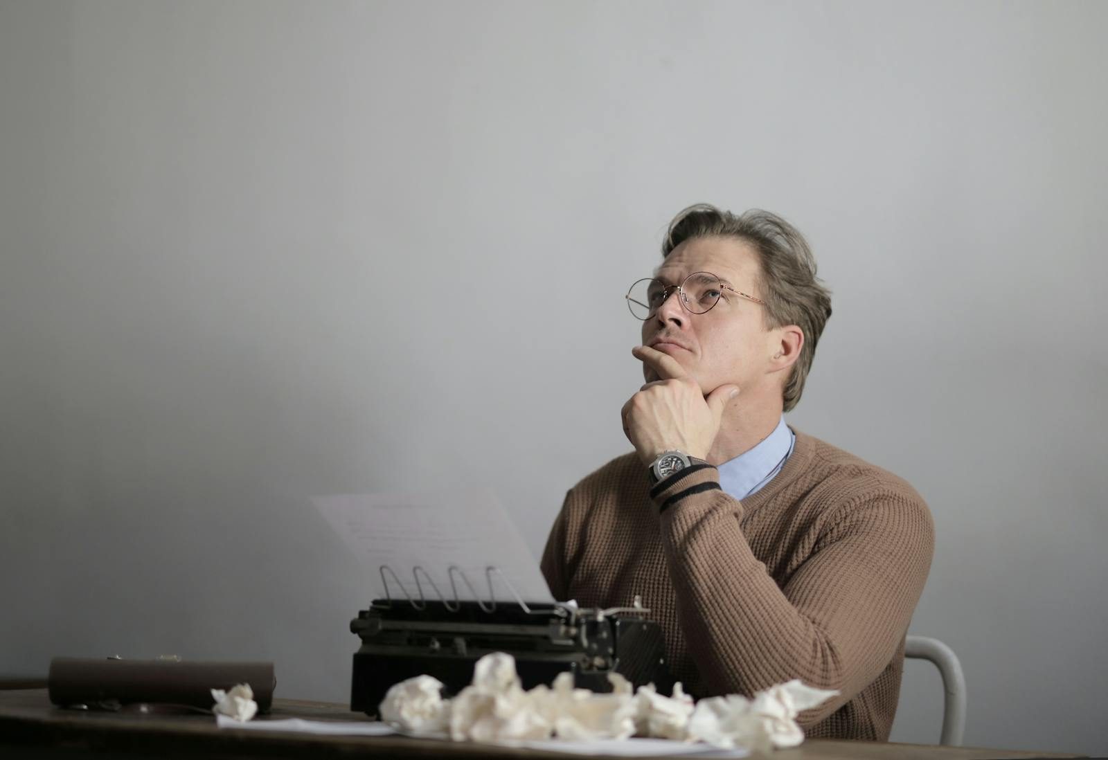 Serious adult casually dressed writer sitting at table using typewriter while pensively with hand at chin looking up and searching for inspiration