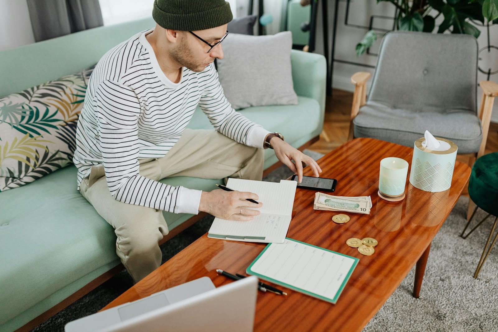 A man in casual attire managing finances with a laptop and notebook on a wooden table indoors.