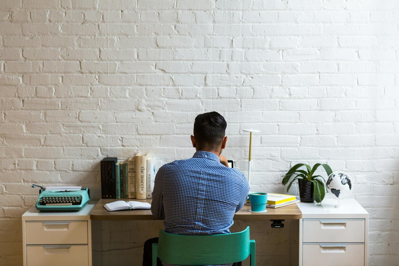 Back view of man at desk with typewriter and books, creating in a minimalist workspace.