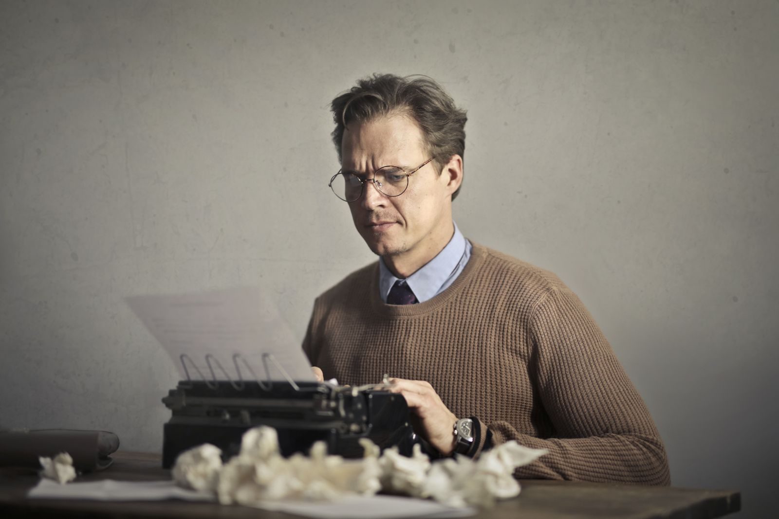 Pensive displeased man in glasses and elegant clothes sitting at table and using typewriter among mush of crumpled sheets of paper while experiencing stress and difficulties in the creative process against gray wall in office