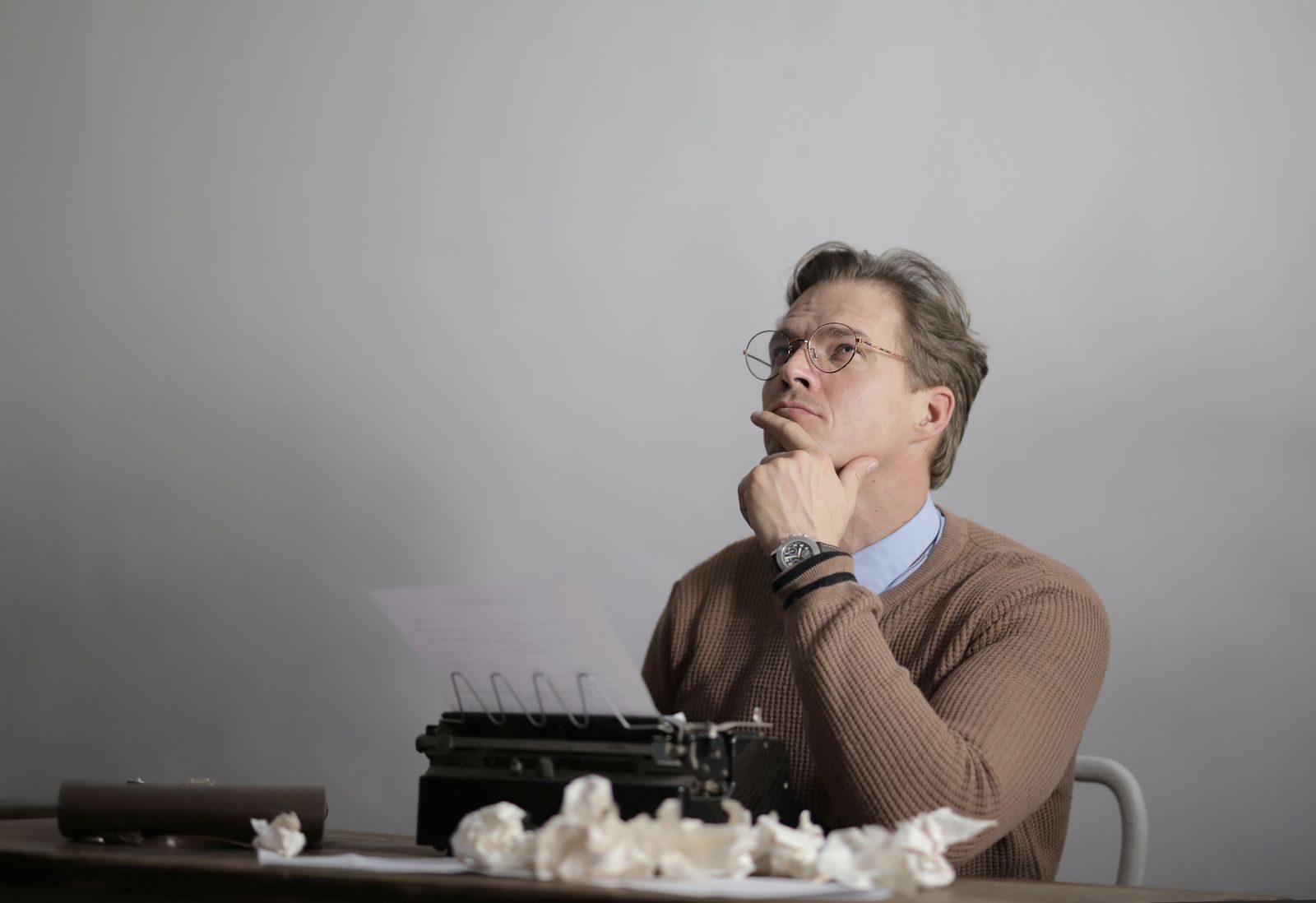 Serious adult casually dressed writer sitting at table using typewriter while pensively with hand at chin looking up and searching for inspiration