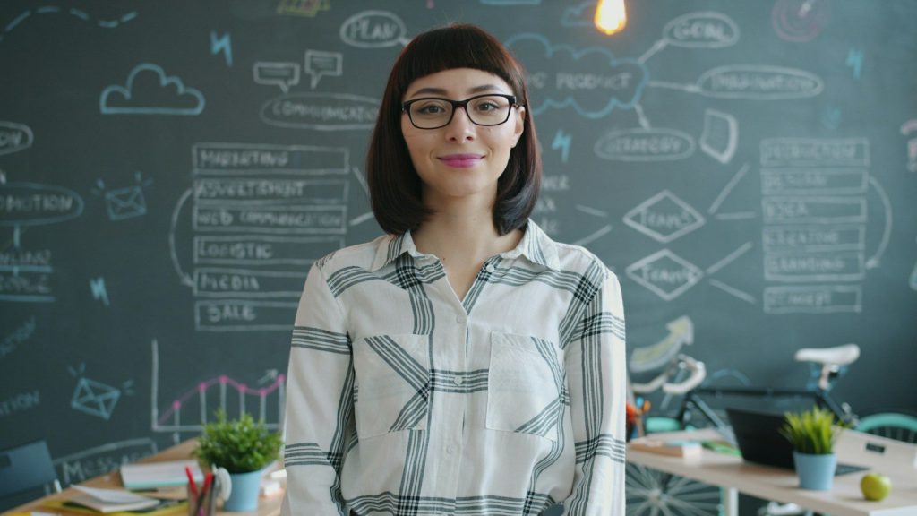 Woman with glasses smiles in front of chalkboard
